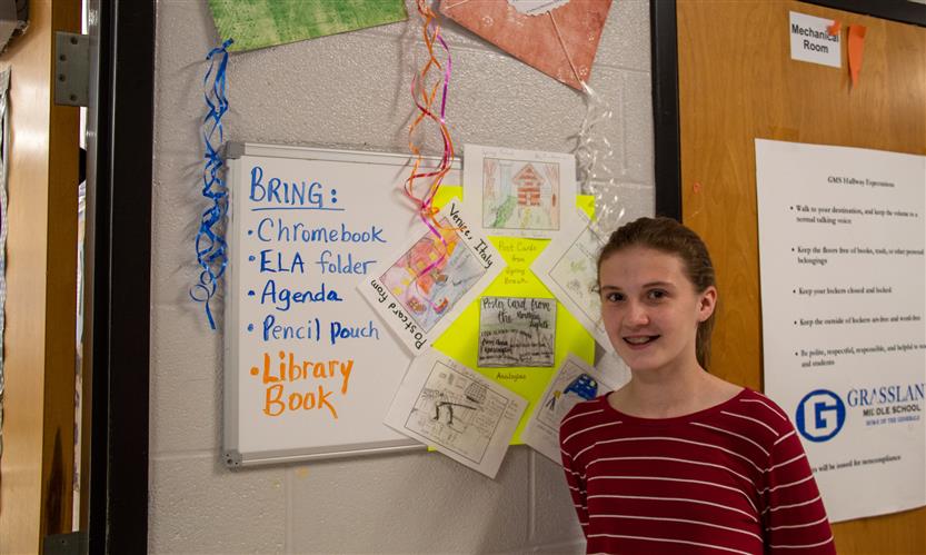A girl with her hair in a ponytail smiles while standing against a wall in a school